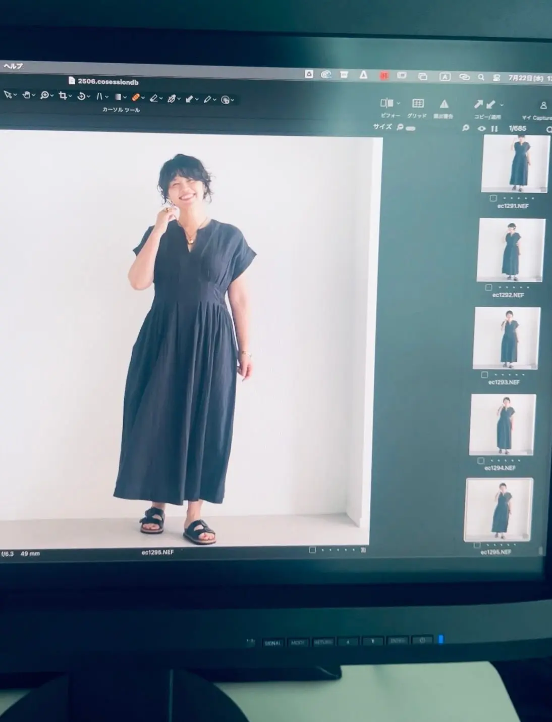 黒いロングワンピースを着た女性が、白い壁の前でポーズをとっている。ワンピースはゆったりとしたシルエットで、茶色のサンダルを履いている。背景には、写真のサムネイルが並んでいる。