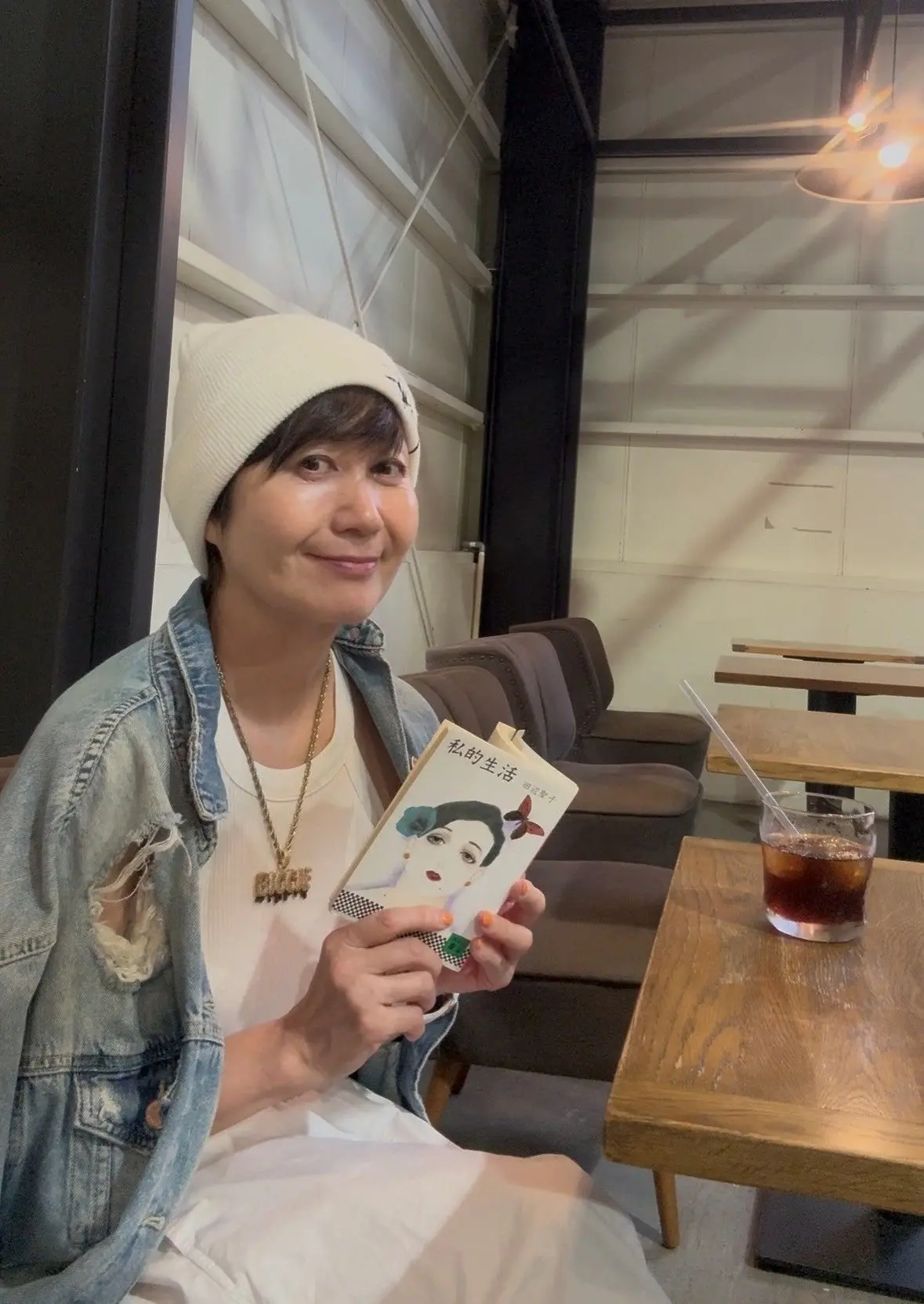ビーニーハットをかぶった女性が、絵入りの本を手に持ってカフェで座っている。木製のテーブルには飲み物が入ったグラスが置かれている。
