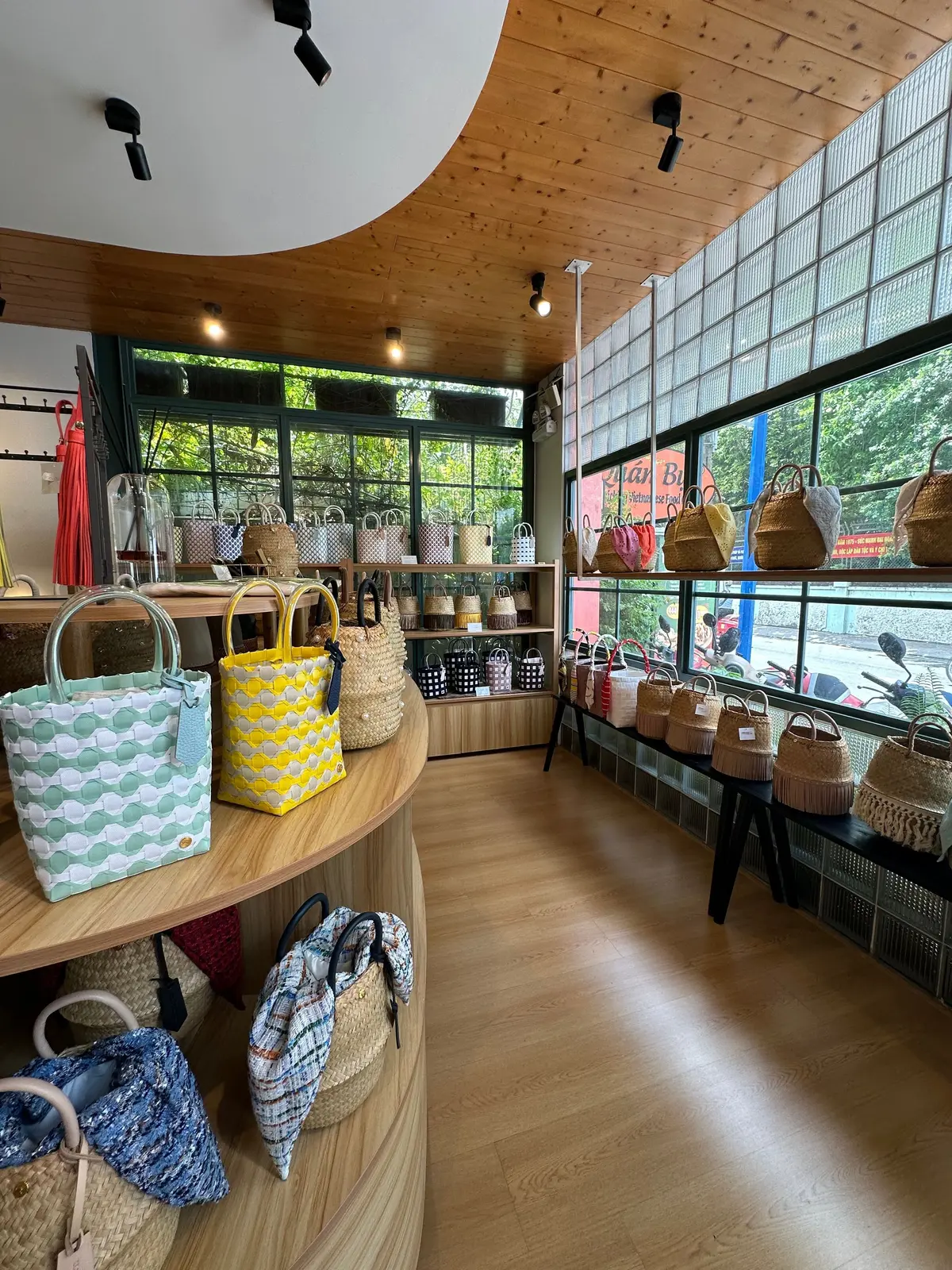 木製の床と天井の明るい店内には、さまざまな色や柄のトートバッグやかごが展示されています。木製の棚やディスプレイ台に並べられ、窓から自然光が差し込んでいます。