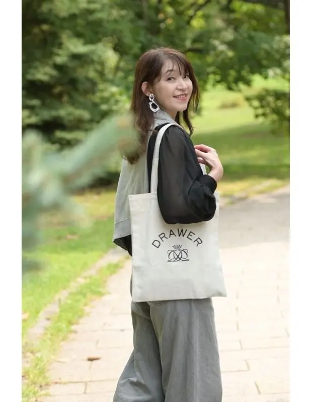 白いトートバッグを持つ女性。グレーのパンツ、黒い長袖ブラウスを着用。背景は緑豊かな公園の遊歩道。右手をバッグの取っ手にかけ、こちらを向いて微笑んでいる。耳には白いイヤリング。