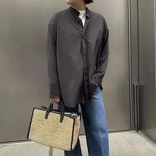 黒いオーバーサイズのシャツにデニムを合わせた女性。白いTシャツを中に着用。手には黒縁の編み込みトートバッグを持っている。背景はグレーの壁。