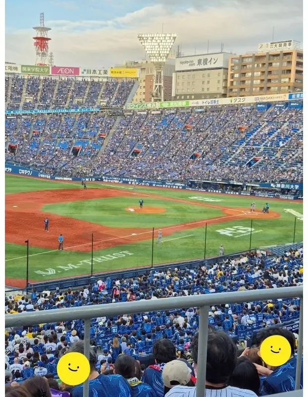 川崎球場での野球試合。満員の観客席が青いユニフォームで埋め尽くされ、グラウンドには選手がいる。背景には高層ビル群と広告看板が見える。