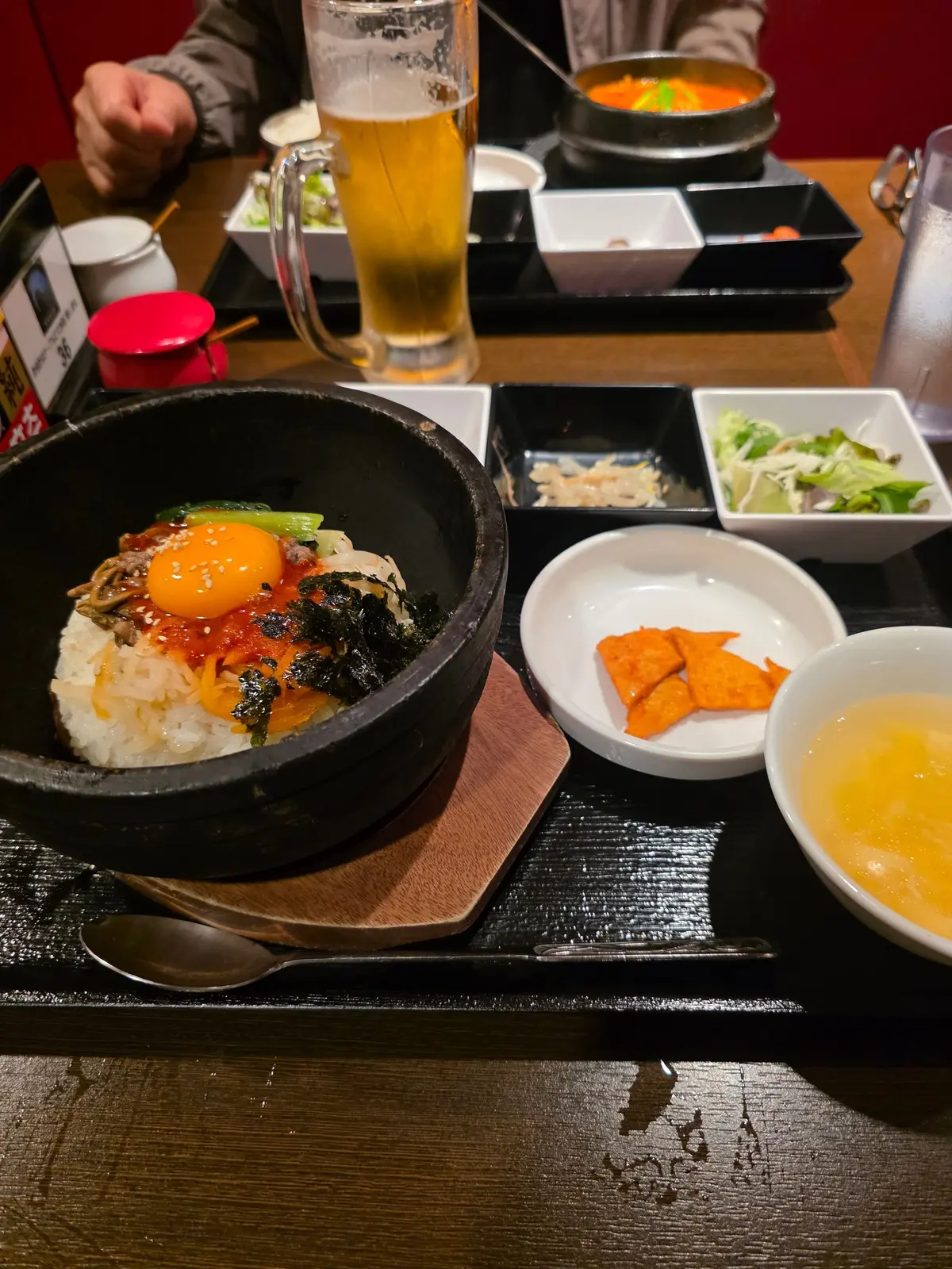 黒い木製のトレイに、石焼ビビンバ、ビール、小皿料理が並んでいる様子。石焼ビビンバは、ご飯の上に野菜、肉、卵、海苔などが乗せられ、中央にコチュジャンが置かれている。ビールのグラスは泡立ち、小皿にはキムチ、オレンジ色の食べ物、スープが入っている。