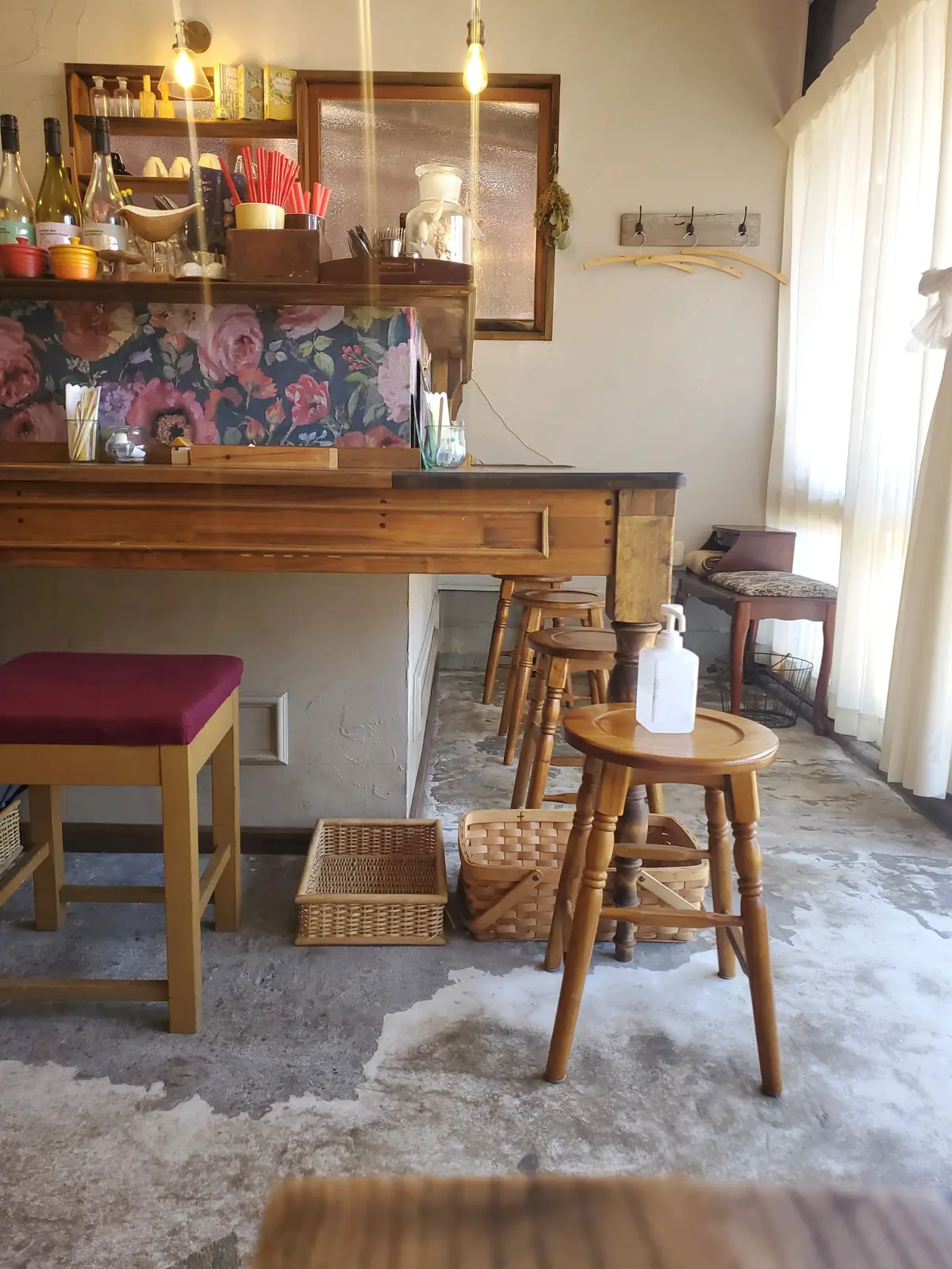 木製のカウンターと背もたれ付きの椅子が並ぶカフェの内部。カウンターは花柄の壁紙で装飾され、上部には照明や調味料が置かれている。床には白いラグが敷かれ、窓からは自然光が差し込んでいる。