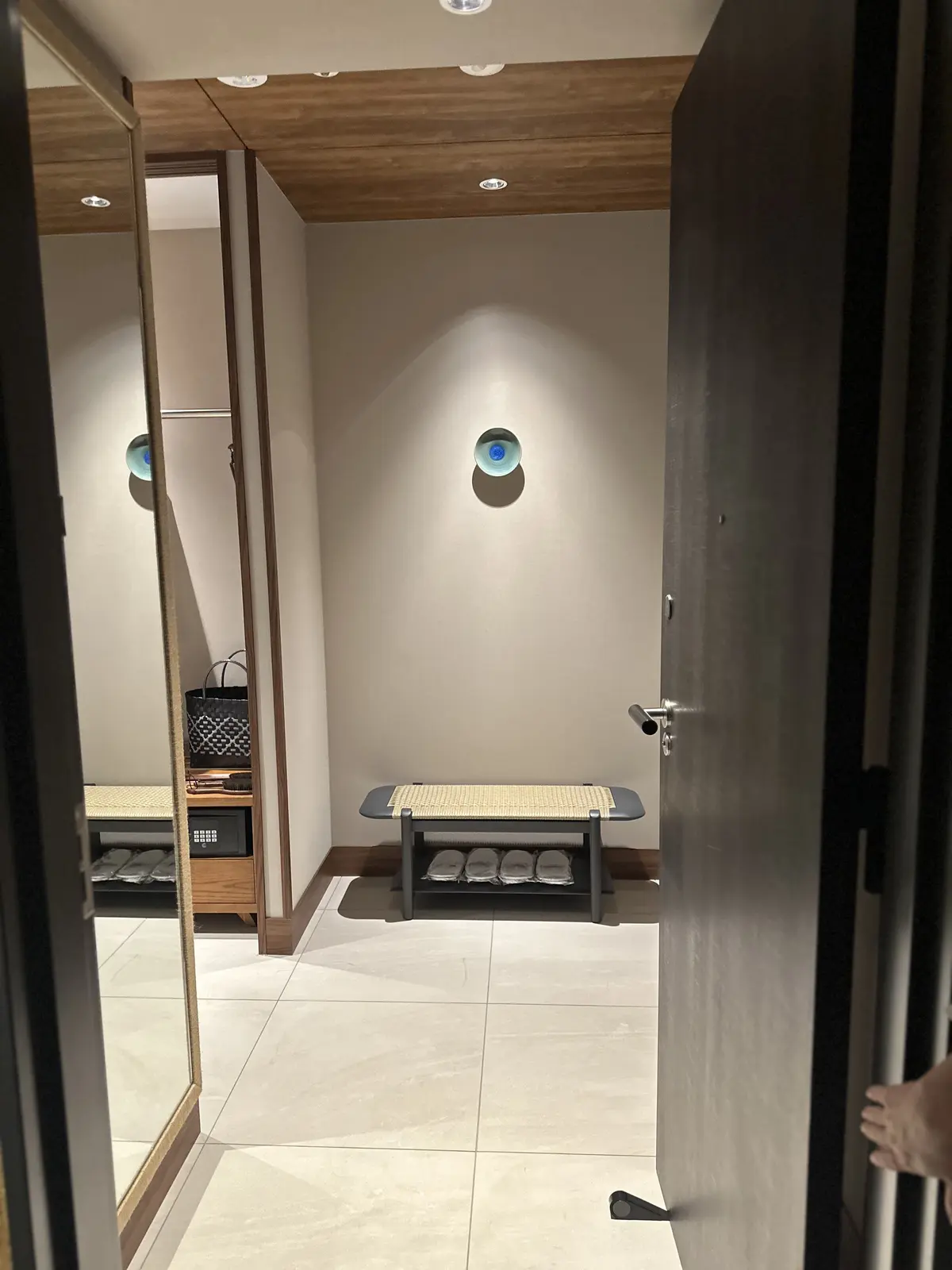試着室の内部。右側に木製の扉が開いており、正面にはベンチと丸い壁掛けの装飾がある。左側には鏡があり、バッグや服が映っている。床は白いタイル張り。
