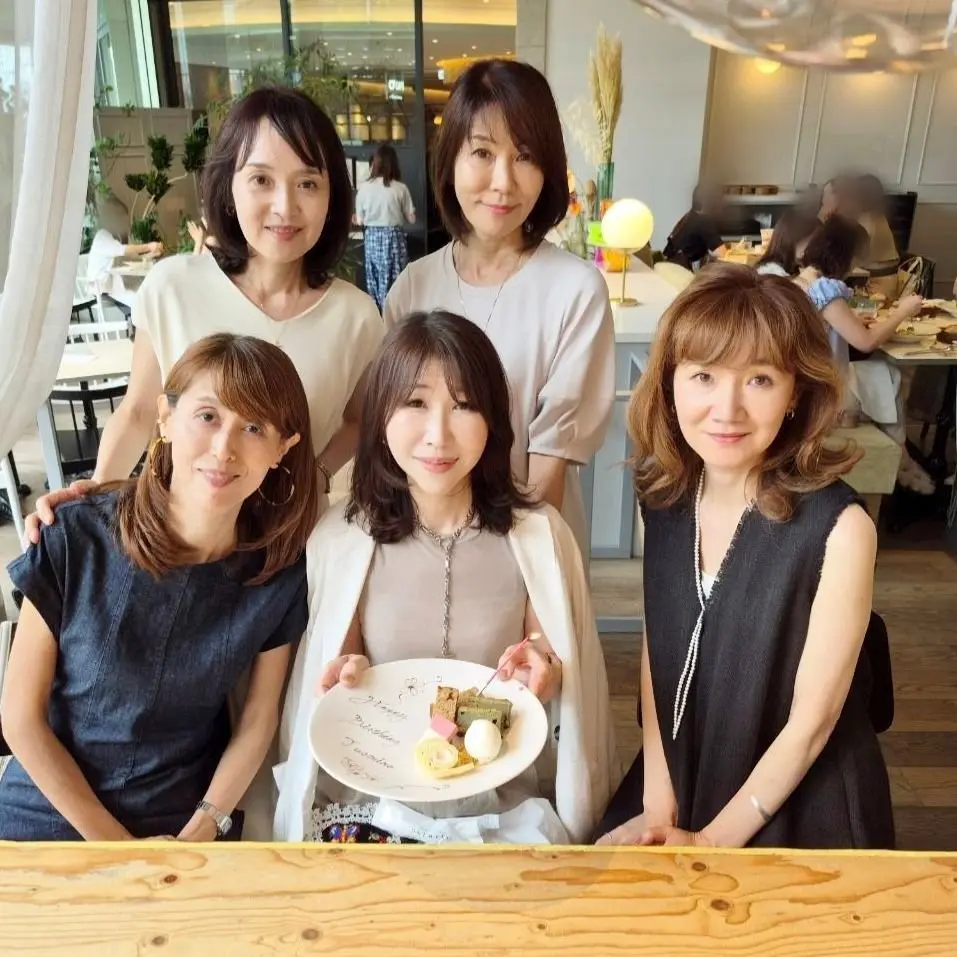 5人の女性がケーキを持って笑顔で写っている写真。左から紺色のワンピースを着た女性、緑色のシャツを着た女性、クリーム色のワンピースを着た中央の女性、黒いワンピースを着た女性、茶色の髪の女性。背景には明るいカフェの様子が見える。