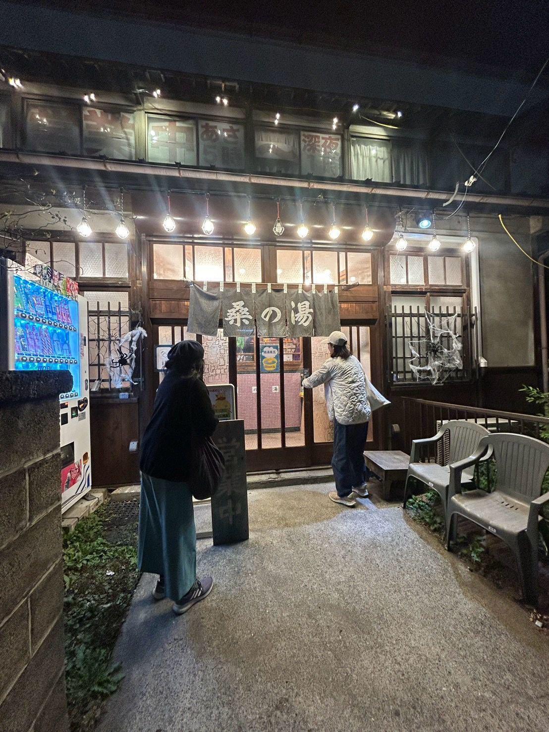 夜の街並み。木造の建物の入り口前に2人並んで立っている。入り口には提灯と看板があり、日本語の文字が書かれている。照明が灯り、温かい光が放たれている。