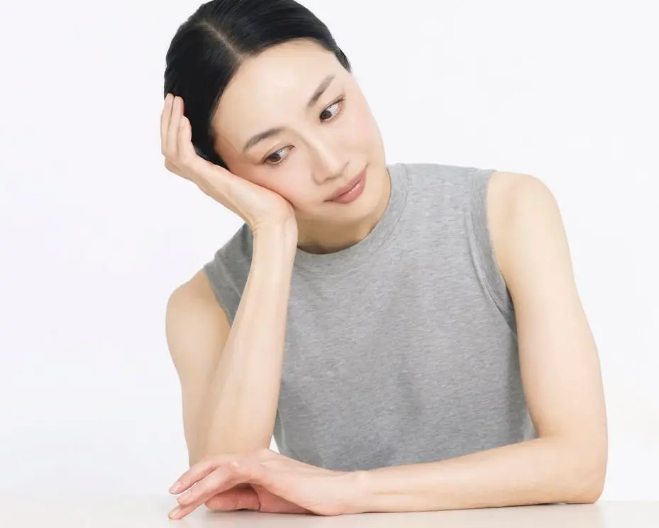 女性がグレーのタンクトップを着て、頭に手を当てて白い背景に座っている