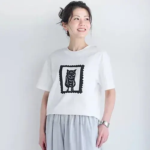 白いTシャツを着た女性が立っている。Tシャツには、黒い猫が描かれたレトロな切手風のデザインがプリントされている。女性は右手を下ろし、微笑んでいる。背景は無地。