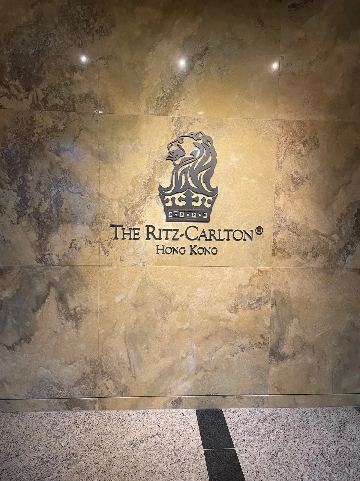 The Ritz-Carlton Hong Kongのロゴが、模様の入ったベージュ色のタイル壁に埋め込まれている。壁の下部にはグレーの帯状の装飾がある。