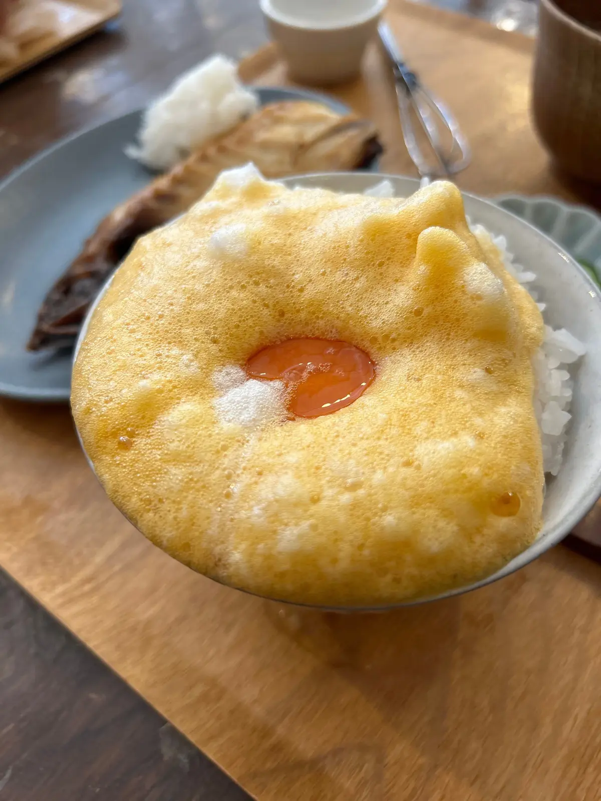 茶色い木製のテーブルに置かれた、ご飯の上に卵焼きが乗ったどんぶり。卵焼きはふっくらとしており、中央に赤い具材が見える。背景には、他の料理と飲み物が置かれたお皿がぼやけて写っている。