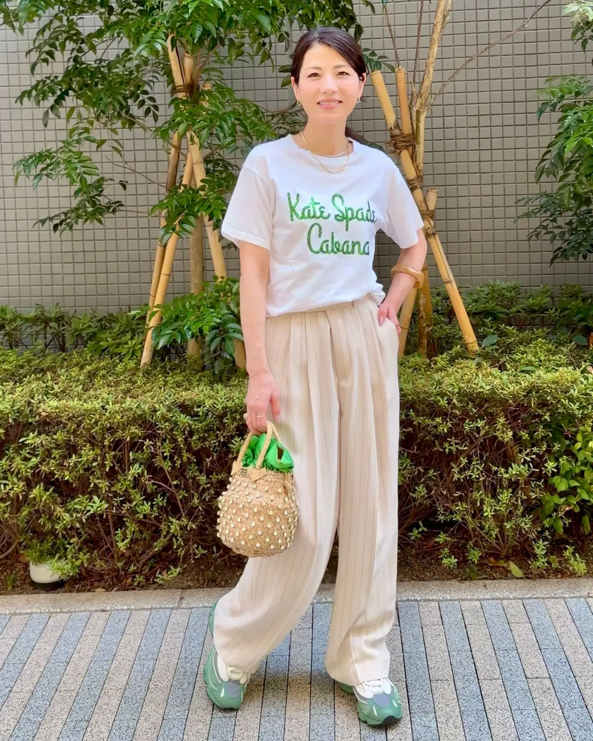 白のTシャツにベージュのパンツを合わせた女性。Tシャツには「Kate Spade Cabana」の刺繍入り。右手に緑色のバッグとストロー製のカゴバッグを持つ。背景には緑の植栽。