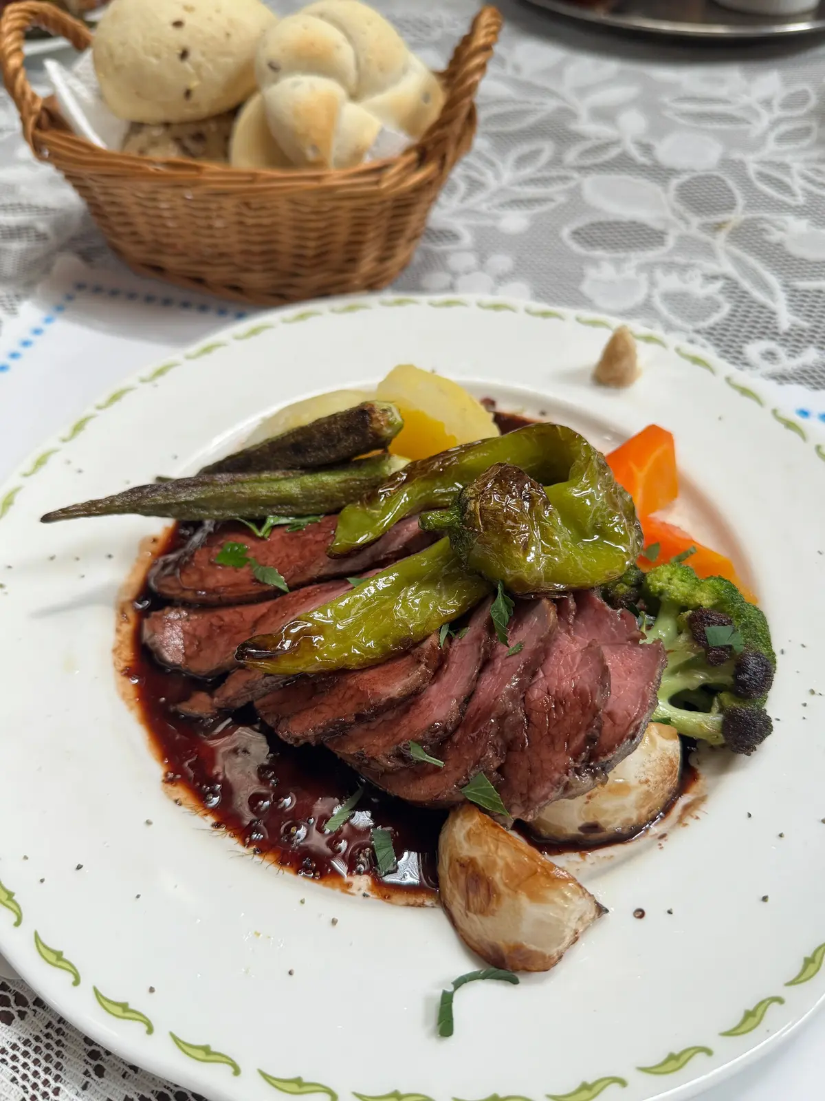 白地に緑色の縁取りの皿に盛り付けられた、牛肉と野菜の料理。牛肉は赤紫色で、緑色のピーマン、黄色いジャガイモ、オレンジ色のニンジン、ブロッコリーなどが添えられている。皿の周りには黒胡椒が散らばり、ソースがかけられている。