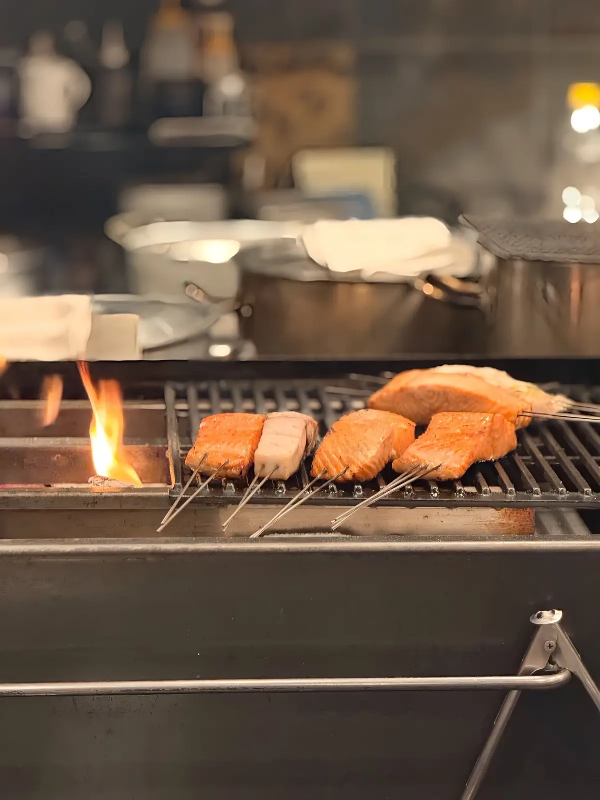 鉄製のグリルで、鮭と豆腐が焼かれている。オレンジ色の鮭の表面に焼き色がつき、豆腐は白く、串が刺さっている。グリルからは炎が上がり、背景には調理器具がぼやけて見える。