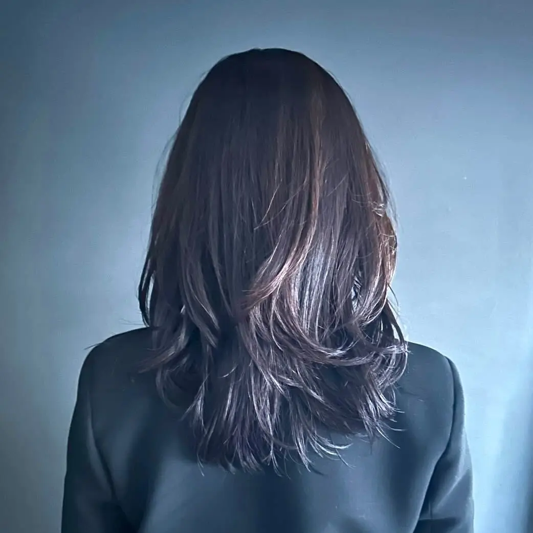 黒いジャケットを着用した人物の後ろ姿。肩から背中にかけて、ダークブラウンの長髪がゆるやかに波打ち、レイヤーカットで軽さを出している。背景はグレーの壁。