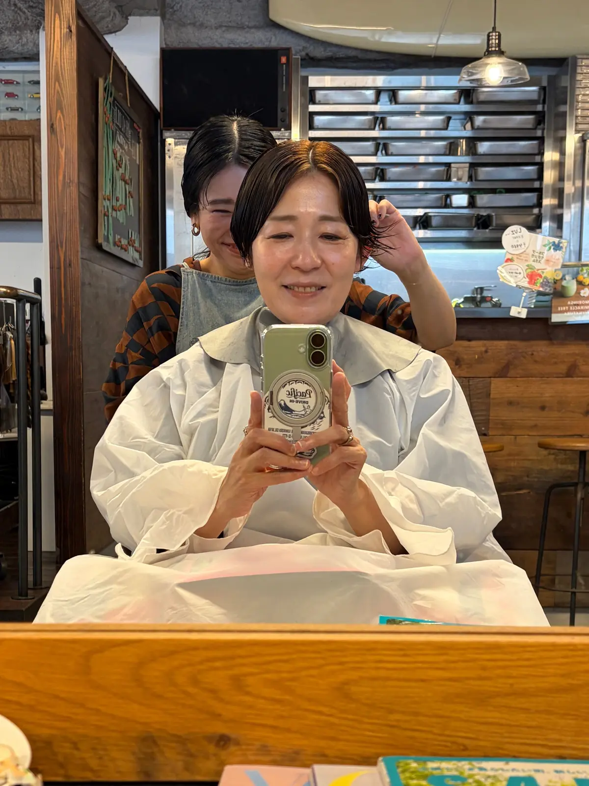 白いシャツを着た女性が自撮りをしている写真。背景には別の女性とカフェのカウンターが見える。女性は笑顔で、スマートフォンを手にしている。