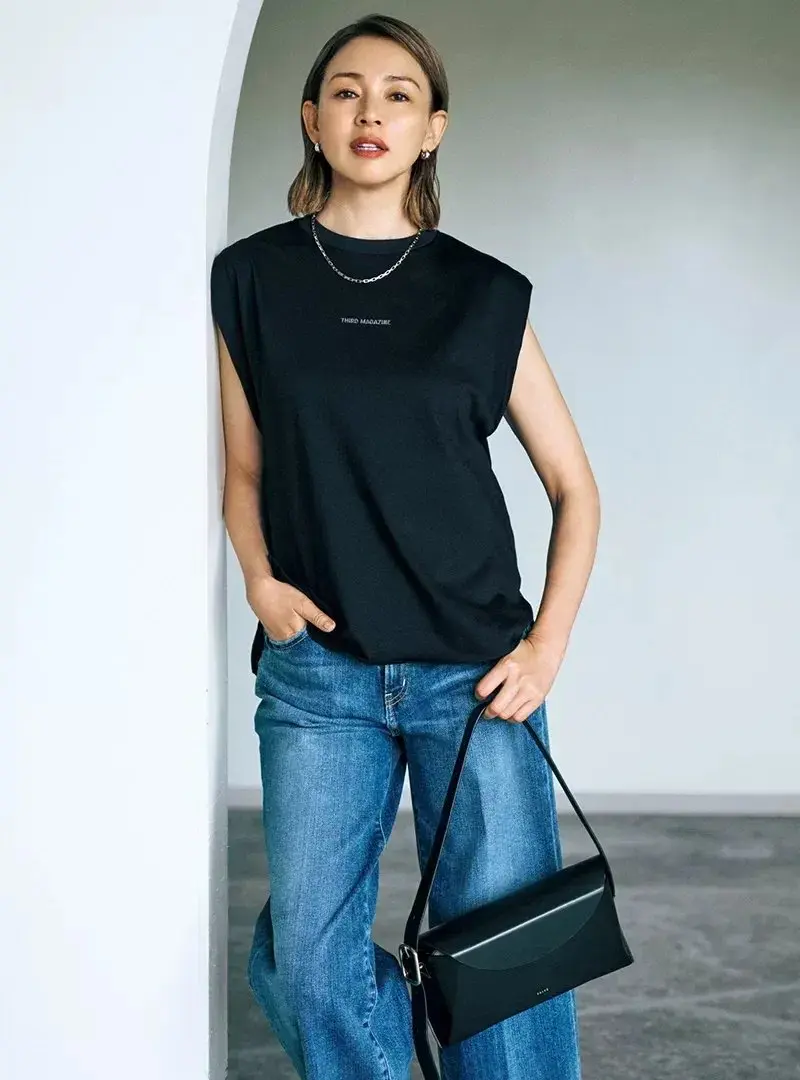 黒いスリーブレスTシャツにデニムを着用した女性。右手に黒いショルダーバッグを持ち、正面を向いている。背景は白いアーチ状の壁。