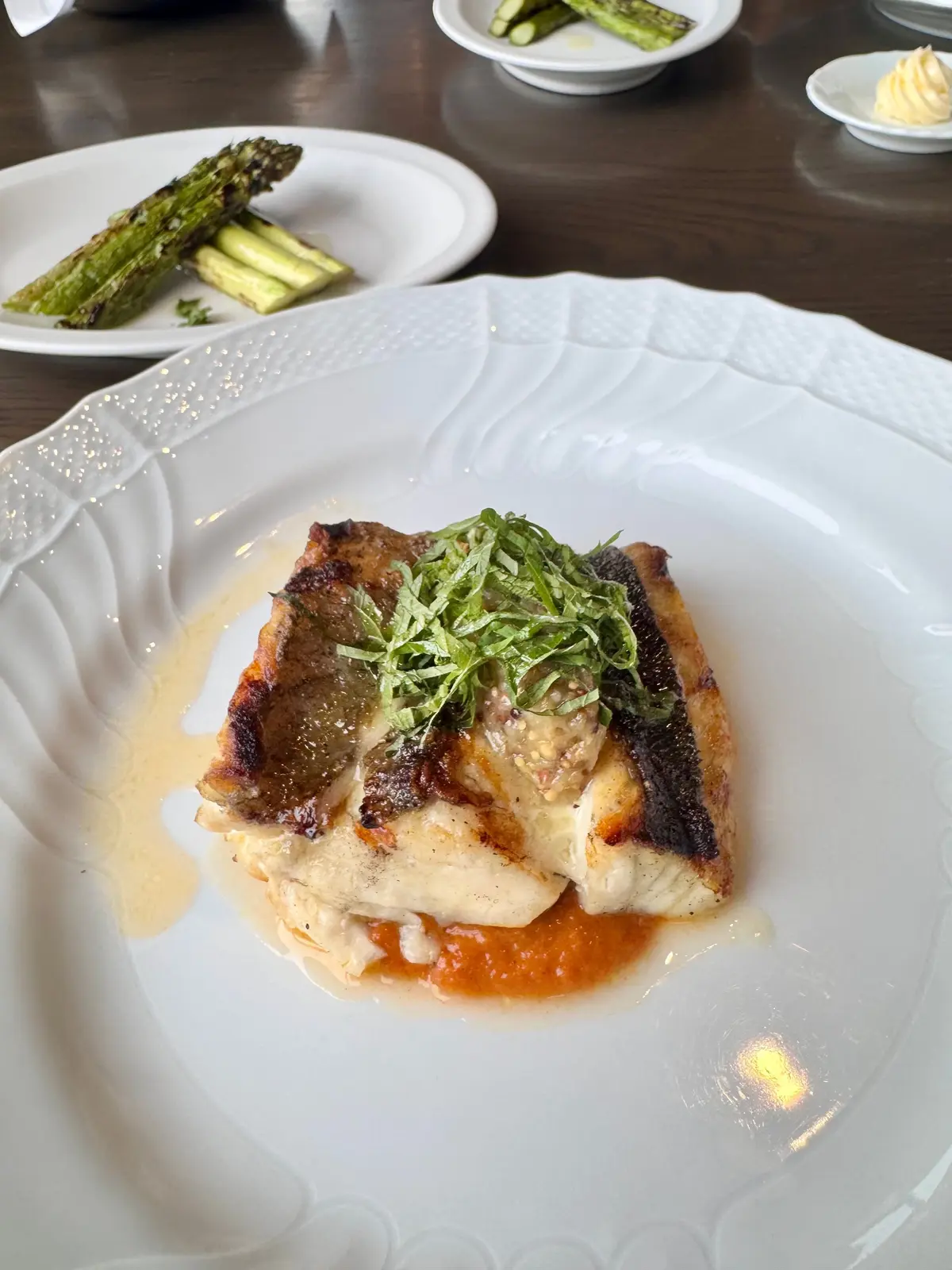 白いプレートに、焼き魚に野菜をトッピングした料理。オレンジ色のソースと緑のハーブが添えられ、背景にアスパラガスとキュウリが並ぶ。