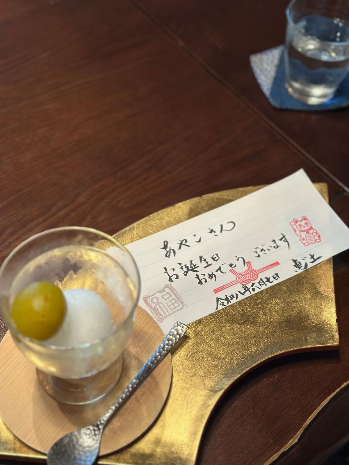 木製のテーブルに置かれた、グラスに入ったレモンとスプーン、そして手書きのメッセージカード。カードには赤い文字で日本語が書かれており、金色の縁取りがある。背景には水を入れたグラスがぼやけて写っている。