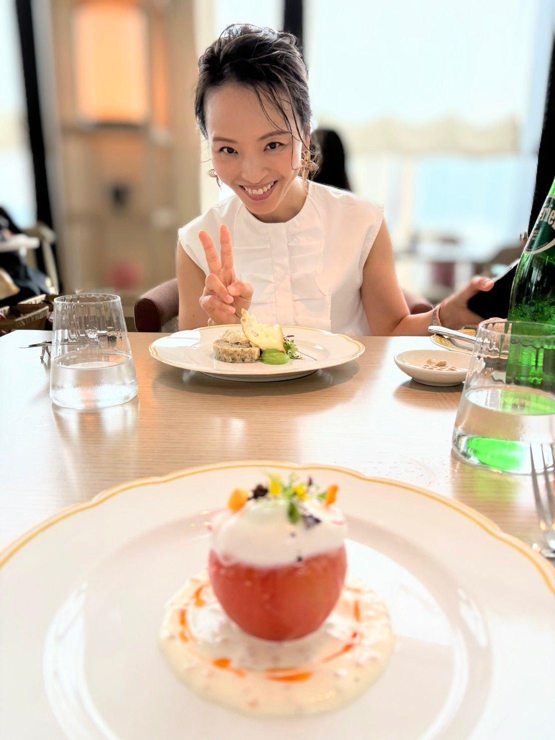 女性が白い服でテーブルに座り、ピースサインを作っている。前に二つの料理が並び、一つは緑の野菜とクラストのある料理、もう一つは赤い果物とクリームのデザート。ガラスの飲み物と小鉢もテーブル上にある。