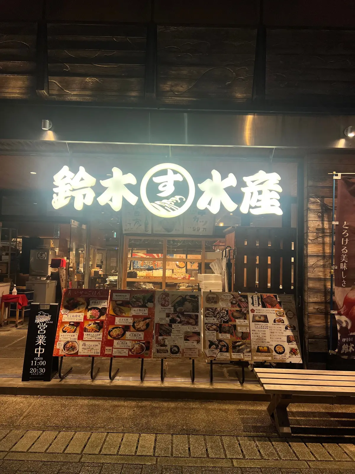 夜の街並みにある、木目調の建物の入り口。看板には「鈴木水産」の文字と丸いロゴマークが浮かび上がる。入り口前にはメニューや広告が立てかけられ、店内は明るく照らされている。