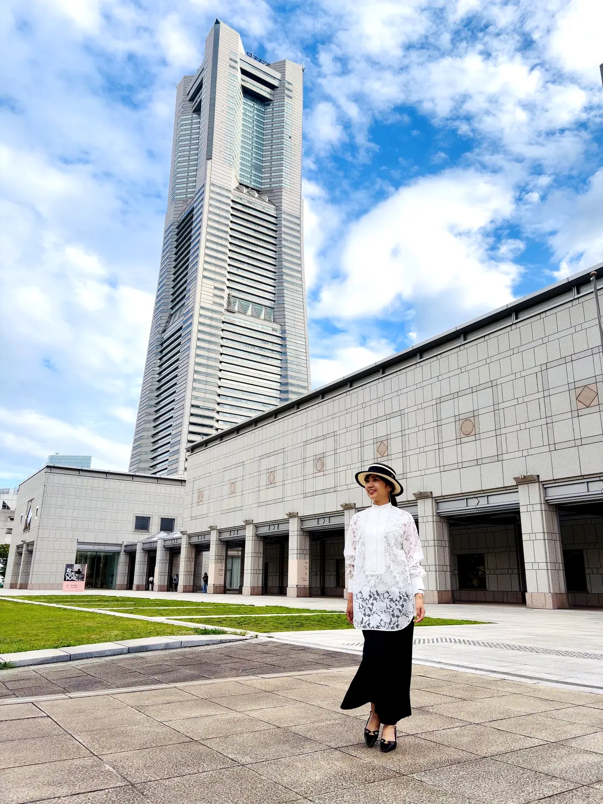 高層ビルと近代的な建物の前で、黒いロングスカートと白いブラウス、帽子をかぶった女性が立っている。青空に白い雲が浮かぶ。