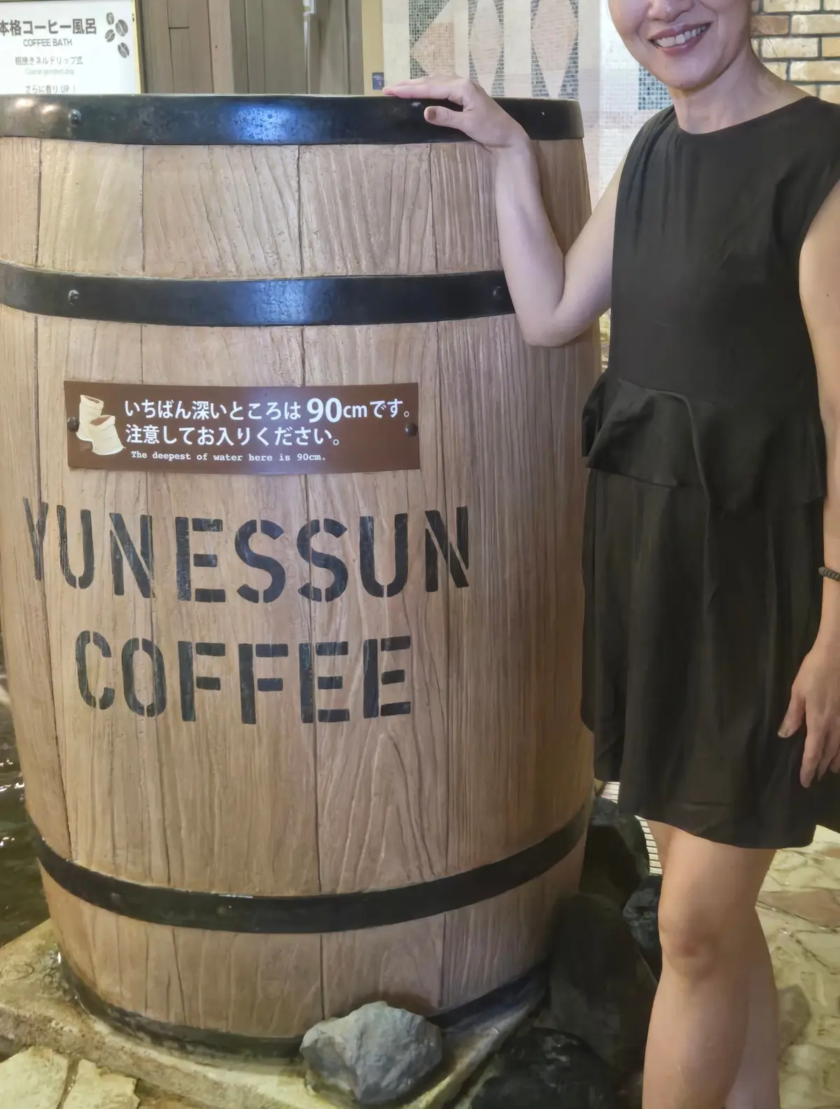笑顔の女性が、巨大な木樽型のコーヒーサーバーの横に立っている。樽には「YUNESSUN COFFEE」の文字が書かれている。背景はレンガの壁で、右にはコーヒーの看板が見える。