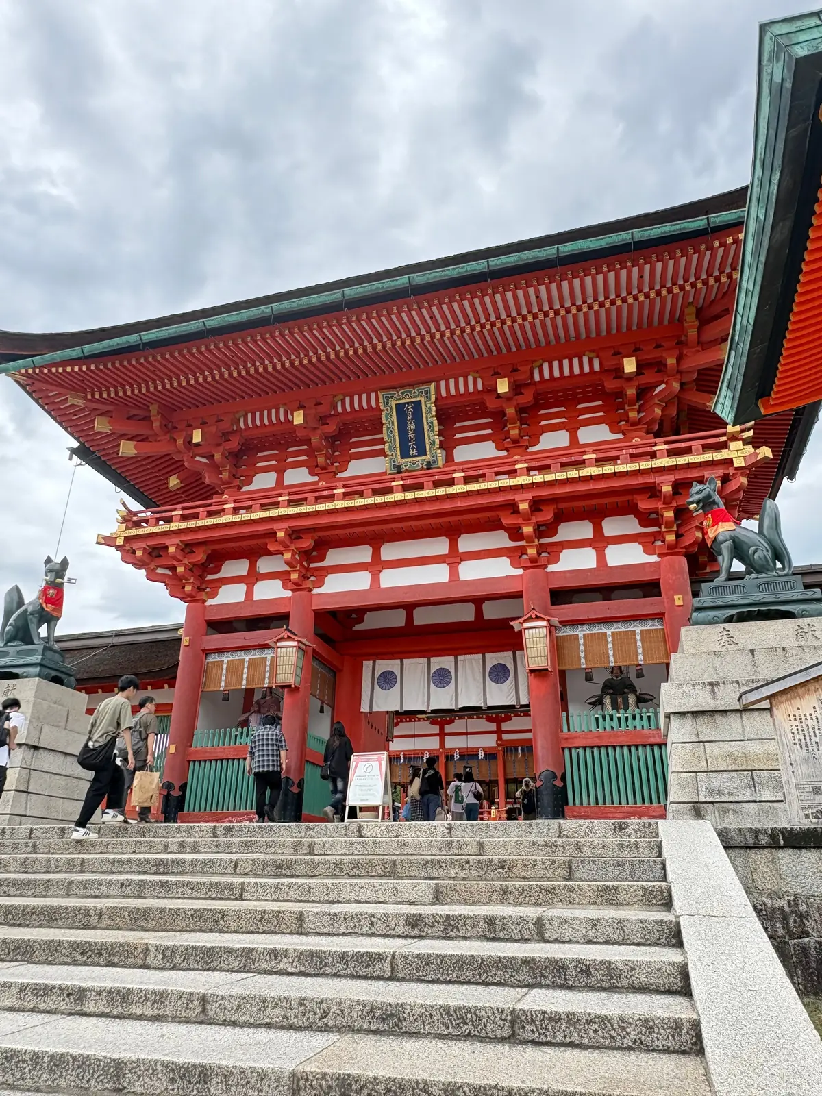 朱色の木造建築が階段の上にそびえ立ち、屋根は緑色。左右には石造りの建物が配置され、石段を人が上がっている。曇り空の下、伝統的な日本の建造物の様子。
