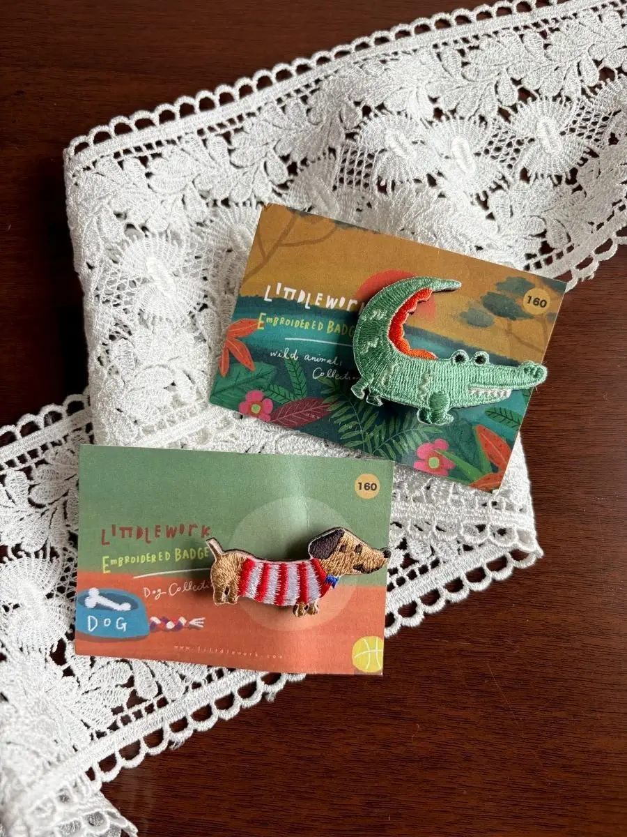 リトルウィークの刺繍バッジ2つ、ワニとドッグが白いレースの上に並ぶ