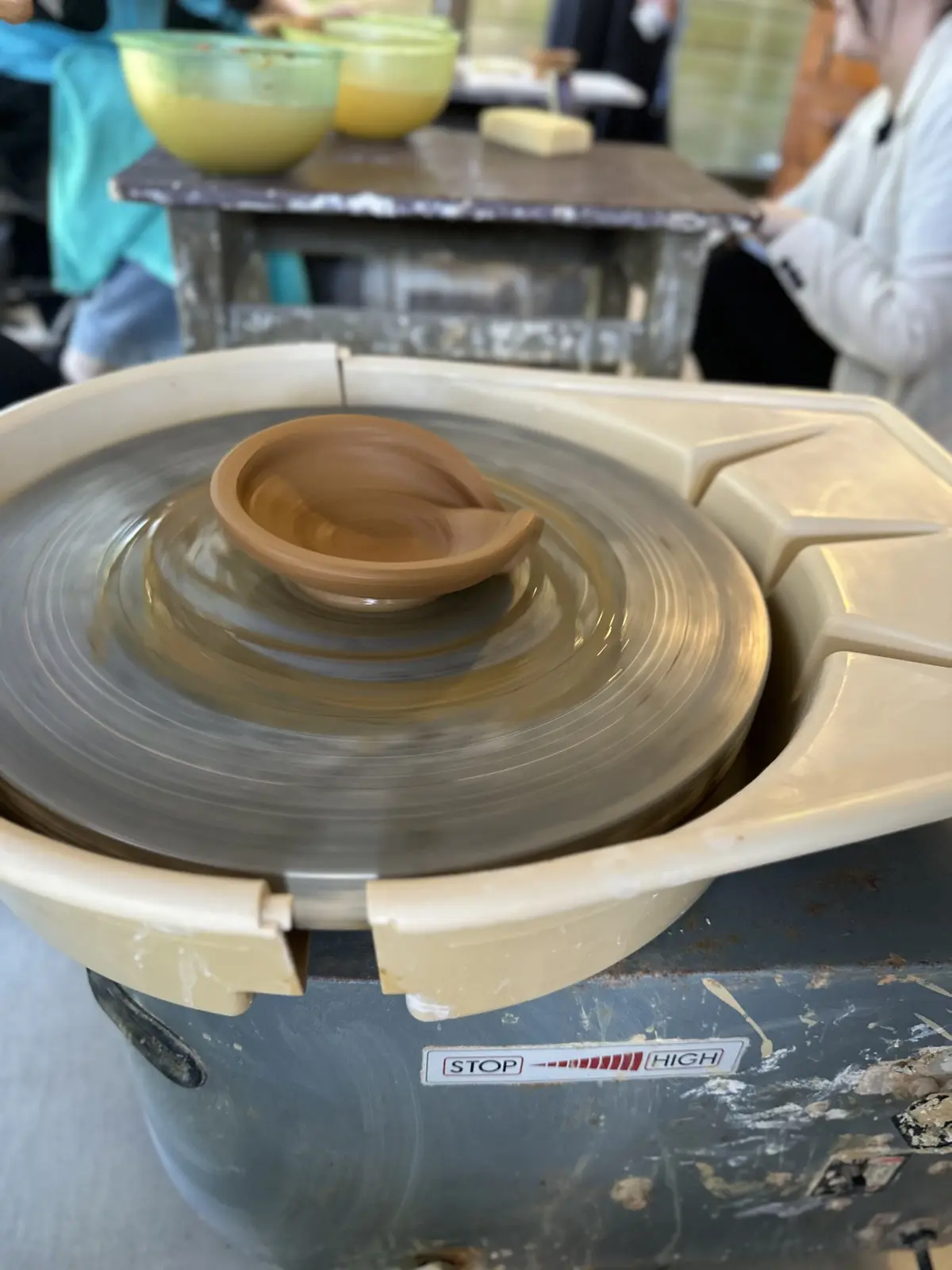 陶芸の電動ろくろで、茶色の粘土でできた作品が回転している。中心部には筒状のものが立ち上がり、周囲に円形の輪がある。背景には作業台と人物が見える。