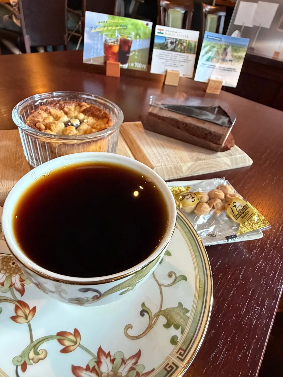 木製のテーブルに置かれた、コーヒー、焼き菓子、おつまみセット。コーヒーは白地の柄入りのカップに入り、焼き菓子は透明な容器、おつまみは木製のプレートに載っている。背景にはメニュー立てがある。