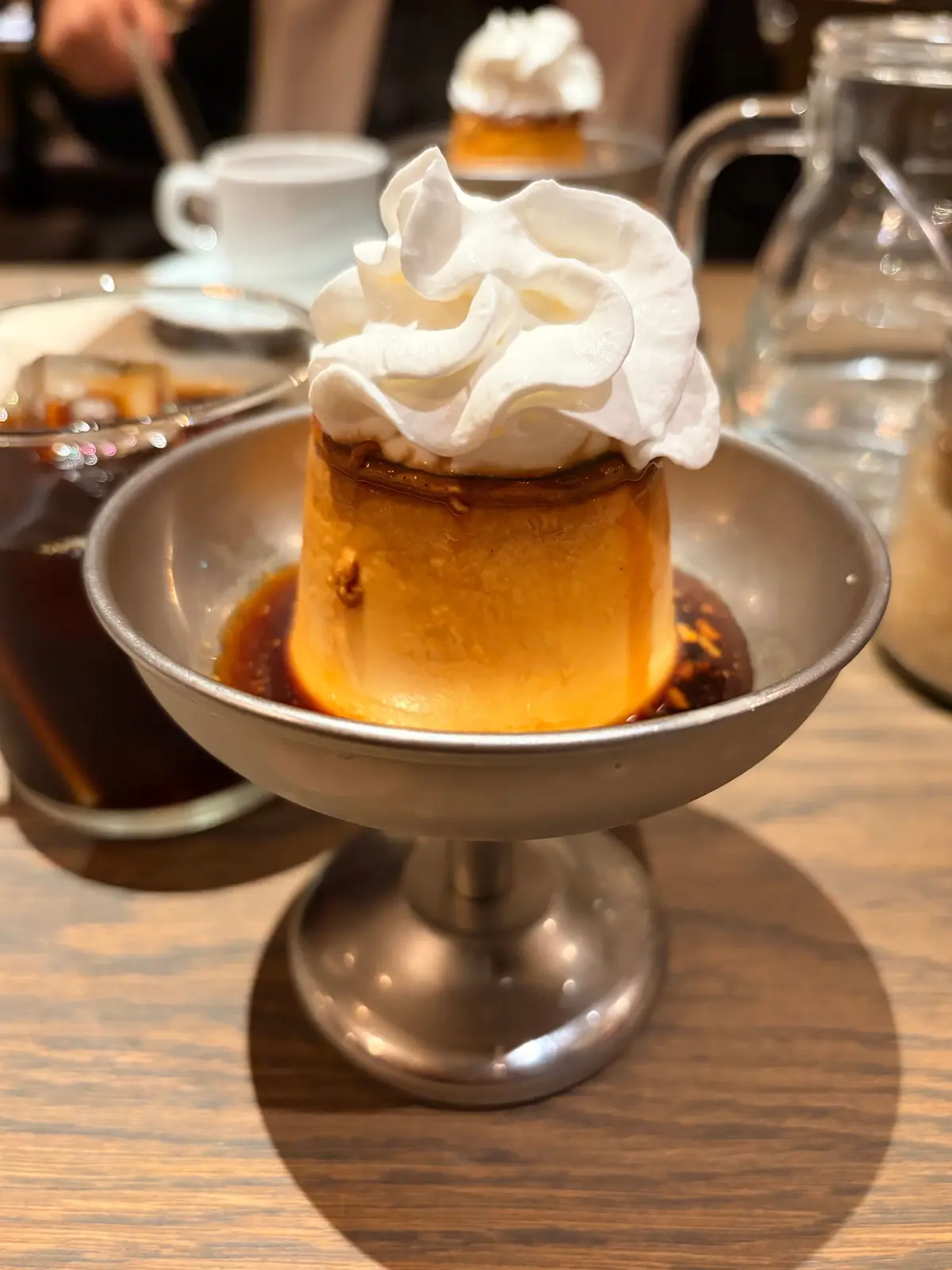 銀色の台に盛り付けられた、カラメルソースをかけたプリン。表面にはたっぷりの生クリームが添えられている。背景にはコーヒーカップやグラスが見える。