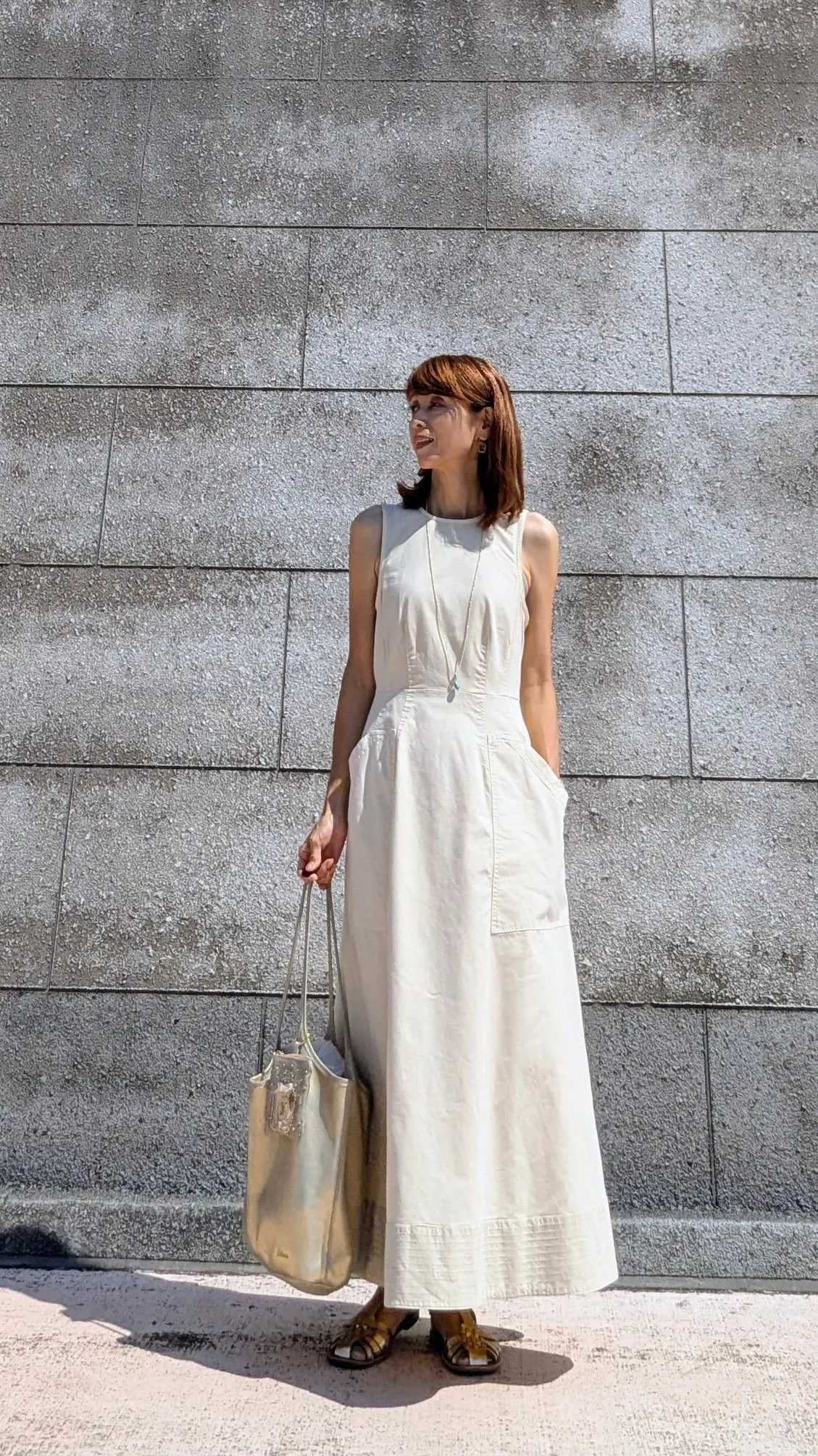 白いロングワンピースを着た女性が、グレーの壁の前に立っている。ゴールドのサンダルを履き、ゴールドのバッグを持ち、シンプルなネックレスをしている。赤みがかった茶色のショートヘア。