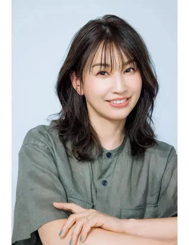 女性が緑色のシャツを着て、腕を組んで微笑んでいる。背景は無地で淡い青色。