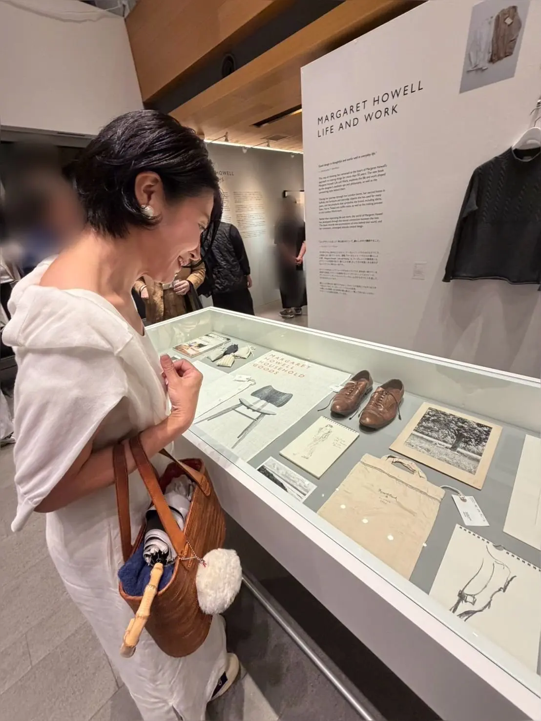 美術館の展示ケースを覗き込む女性。ケース内には、Margaret Howellの文字と、靴、写真、書類などの展示品が並んでいる。女性は茶色のバッグを肩にかけ、白い服を着ている。背景には、Margaret Howellの文字と、黒い服の展示がある。