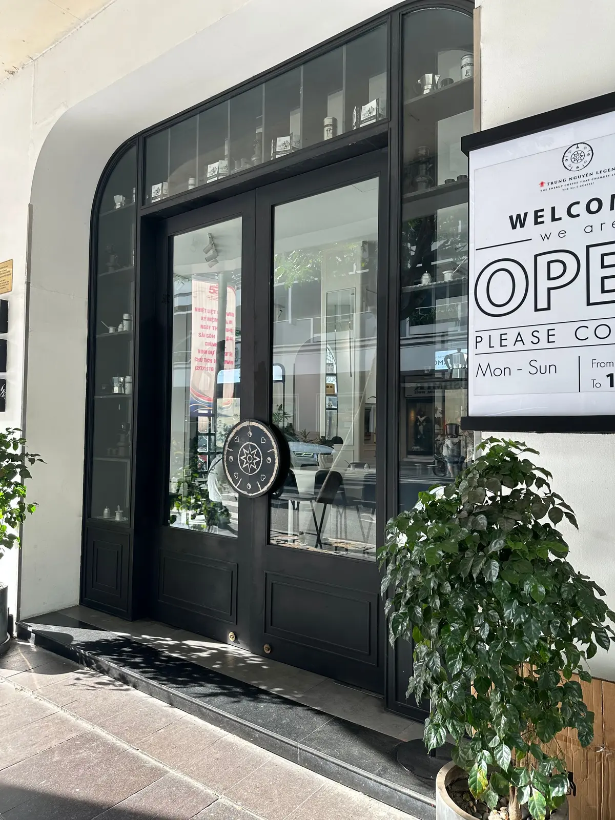ガラスのドアと窓のあるカフェの入口。右側に「OPEN」の看板があり、営業時間も記載されている。ドアの横には緑の観葉植物が置かれている。