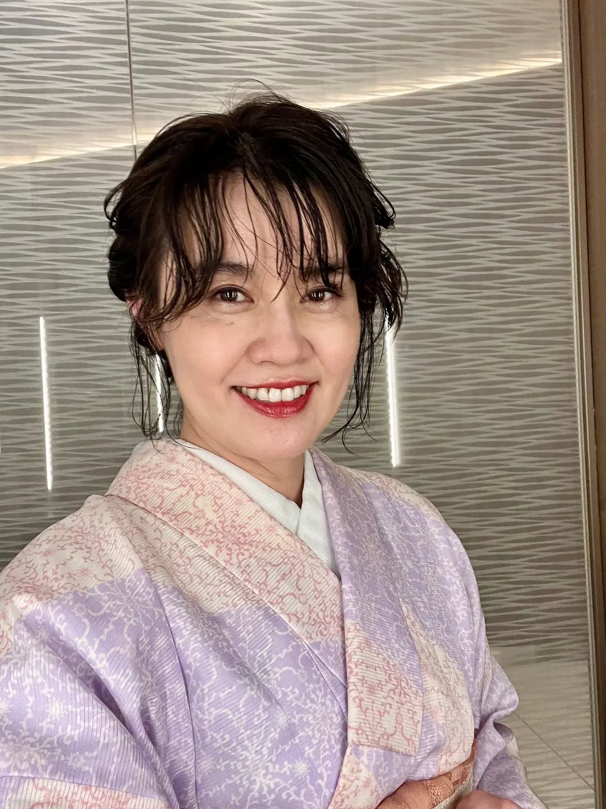 笑顔の女性が、淡い紫色の花柄の着物と白い襦袢を着用してこちらを見ている。黒髪のバングが額にかかり、背景は銀色の波模様の壁。
