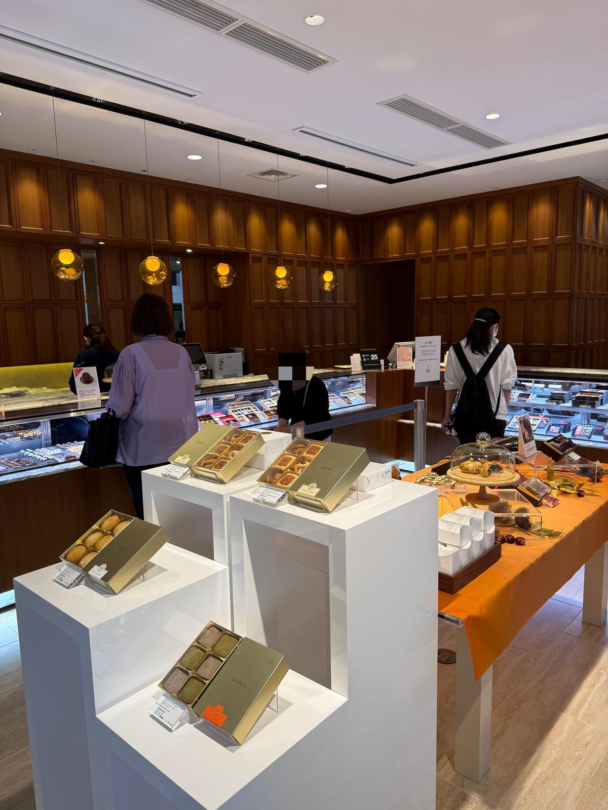 高級チョコレート店「WAKO」の店内。白の台に整列されたチョコレートの箱が並び、奥にはスタッフが対応している様子。木目調の壁と照明が上品な雰囲気を醸し出している。