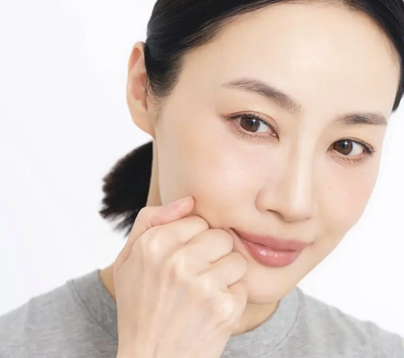 下唇のラインに沿って こぶしを当てて揺らす 黒髪をポニーテールにした女性が、左手で頬を触れ、淡いピンクの唇でほほえんでいる。グレーのタンクトップを着ている。