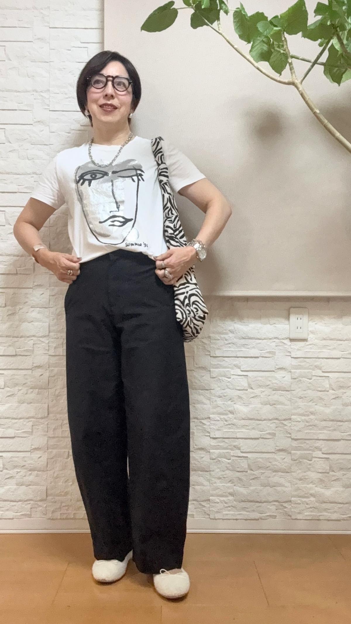 白Tシャツに黒のワイドパンツを合わせた女性。Tシャツには顔のイラストがプリント。黒縁メガネをかけ、ネックレスを着用。右手にゼブラ柄のバッグを持つ。背景には白い壁と観葉植物。