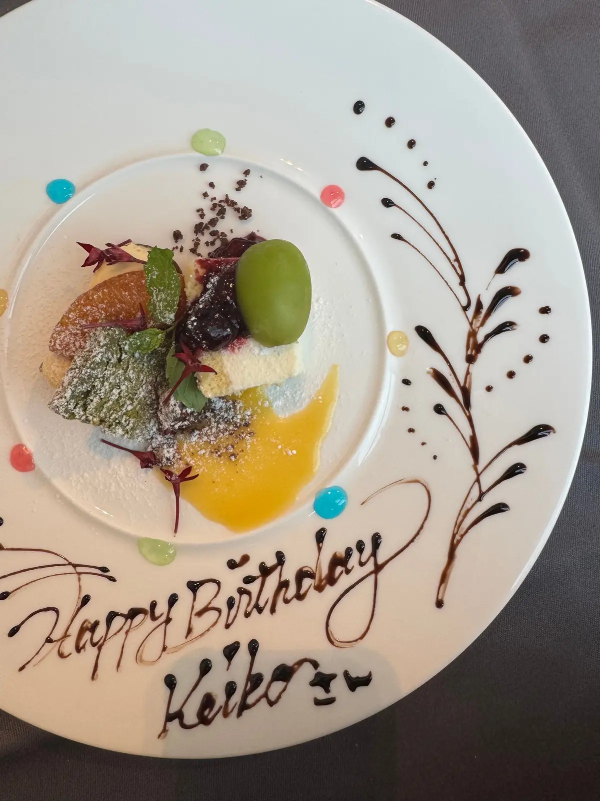 白磁の皿に、チーズケーキとフルーツ、ソースを盛り付けた誕生日デザート。チョコレートソースで「Happy Birthday Keiko」と手書き風にメッセージが描かれている。