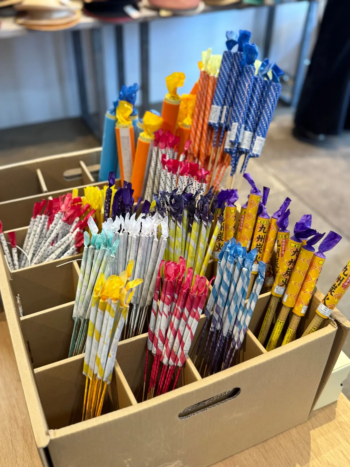 段ボール箱に入った、様々な色と柄の日本製の花火。紫、黄、赤、青など、複数の色合いが混ざり合い、竹串に刺さっている。