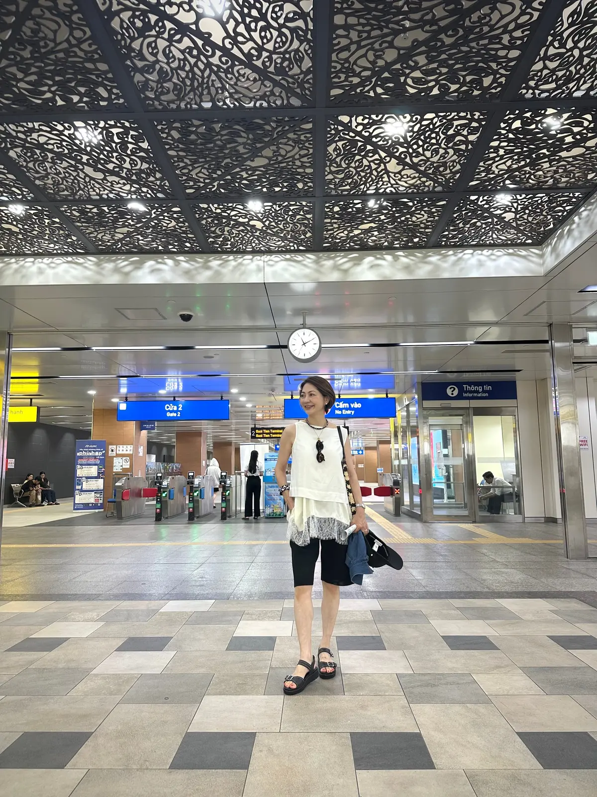 ホーチミンの地下鉄、市民劇場駅の改札前で写真を撮りました。私は白いレースのキャミソールの上に、白いユニクロのノースリーブを重ね着し、黒のハーフパンツを履いています。背景には地下鉄の自動改札と看板、時計があります。