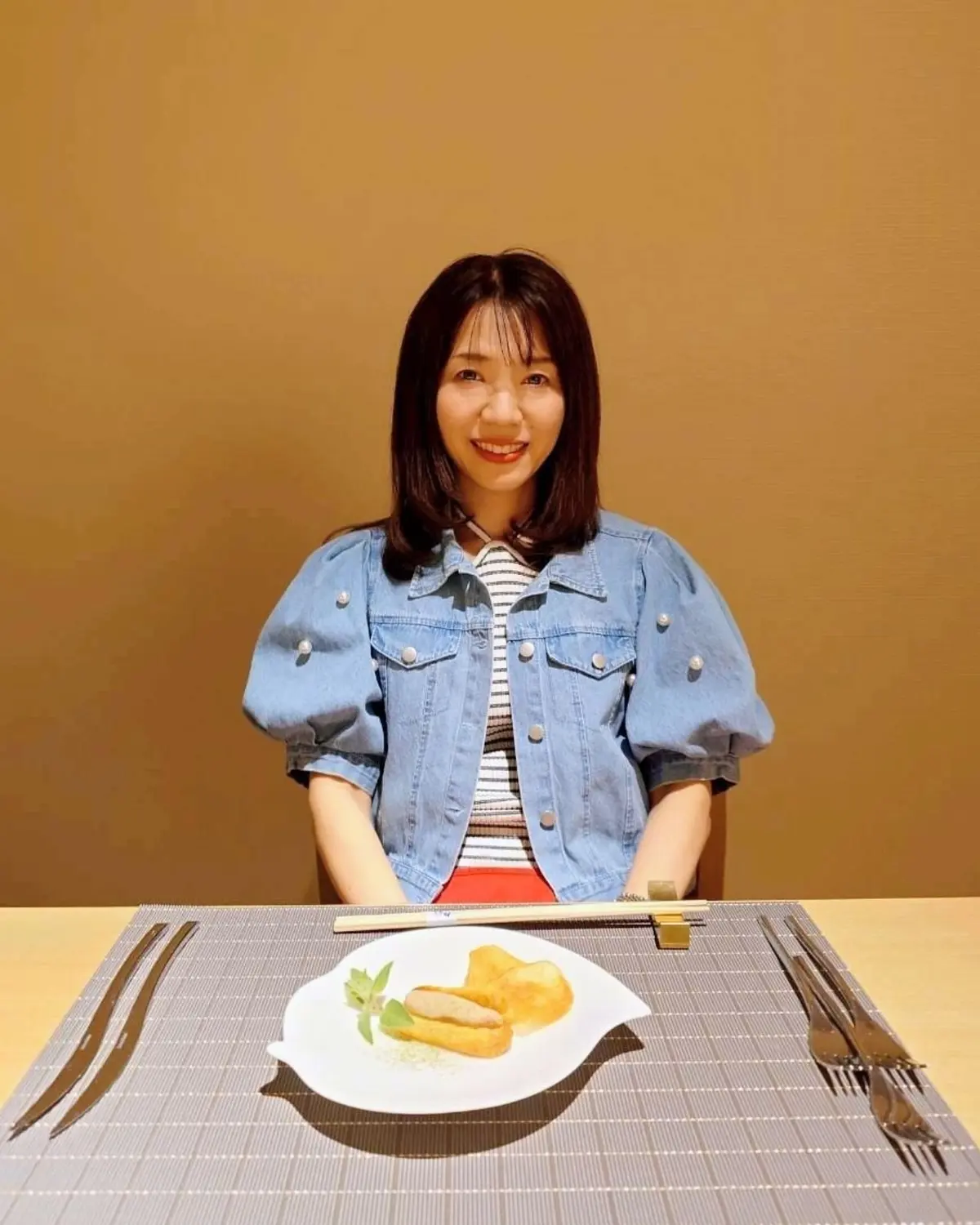 茶色い背景に、女性がデニムジャケットを着て座っている。前に、料理が置かれたプレートと箸、ナイフ、フォークが並んでいる。