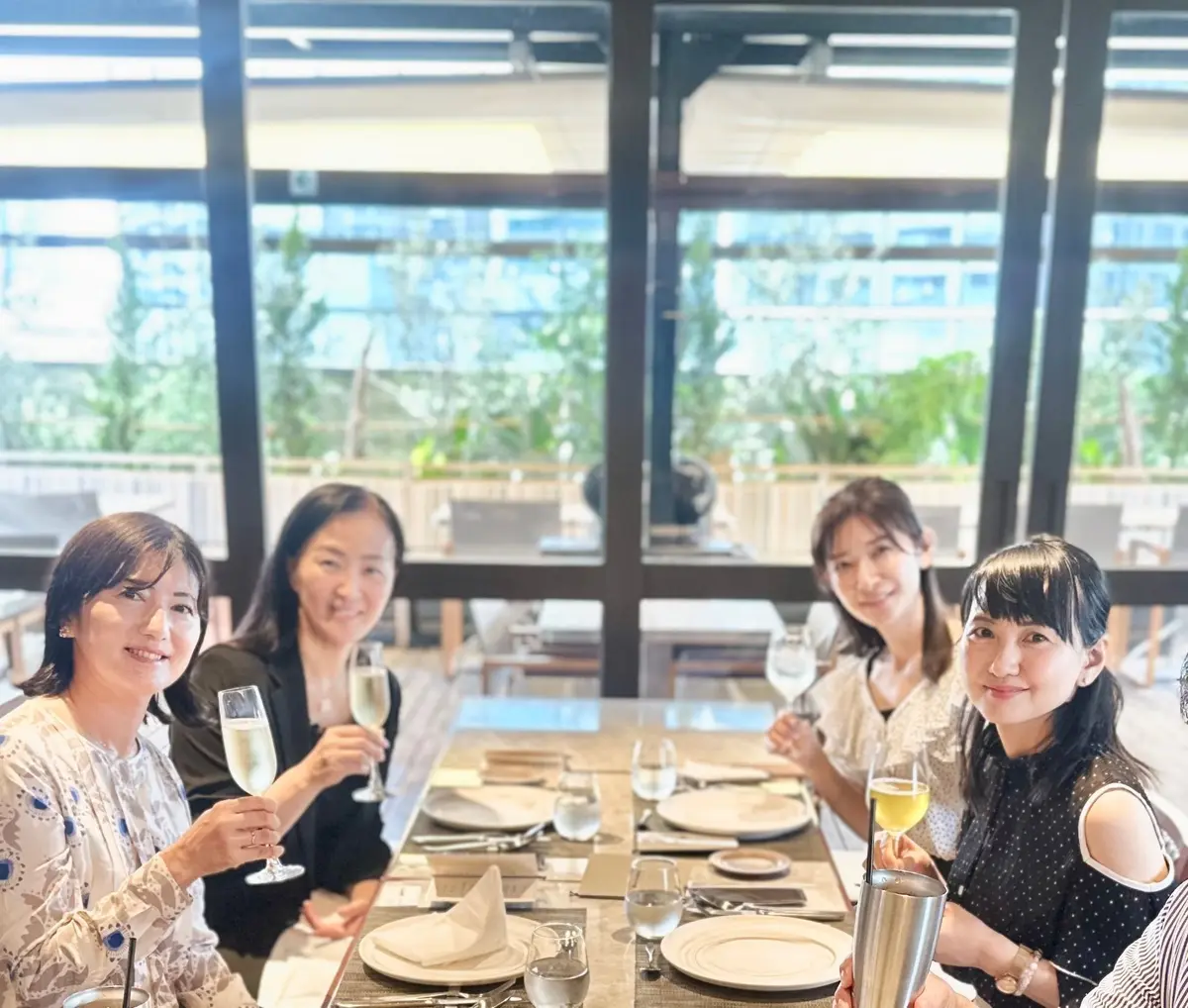 レストランでワイングラスを持つ5人の女性。窓の外には緑豊かな景色が広がり、テーブルには食器が並んでいる。