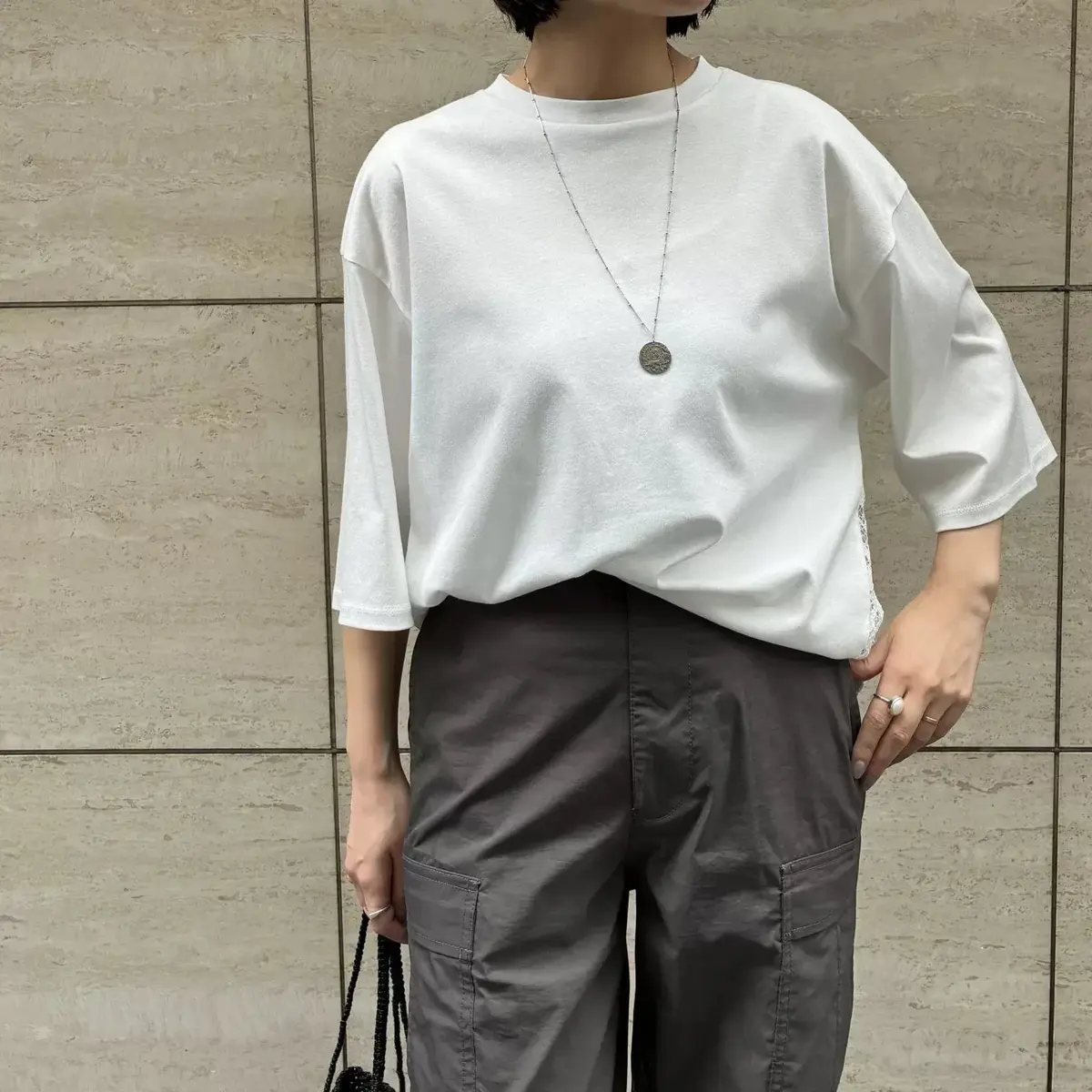 白いオーバーサイズのTシャツとグレーのカーゴパンツを着用した女性。シルバーのネックレスを身につけ、背景はベージュのタイル壁。