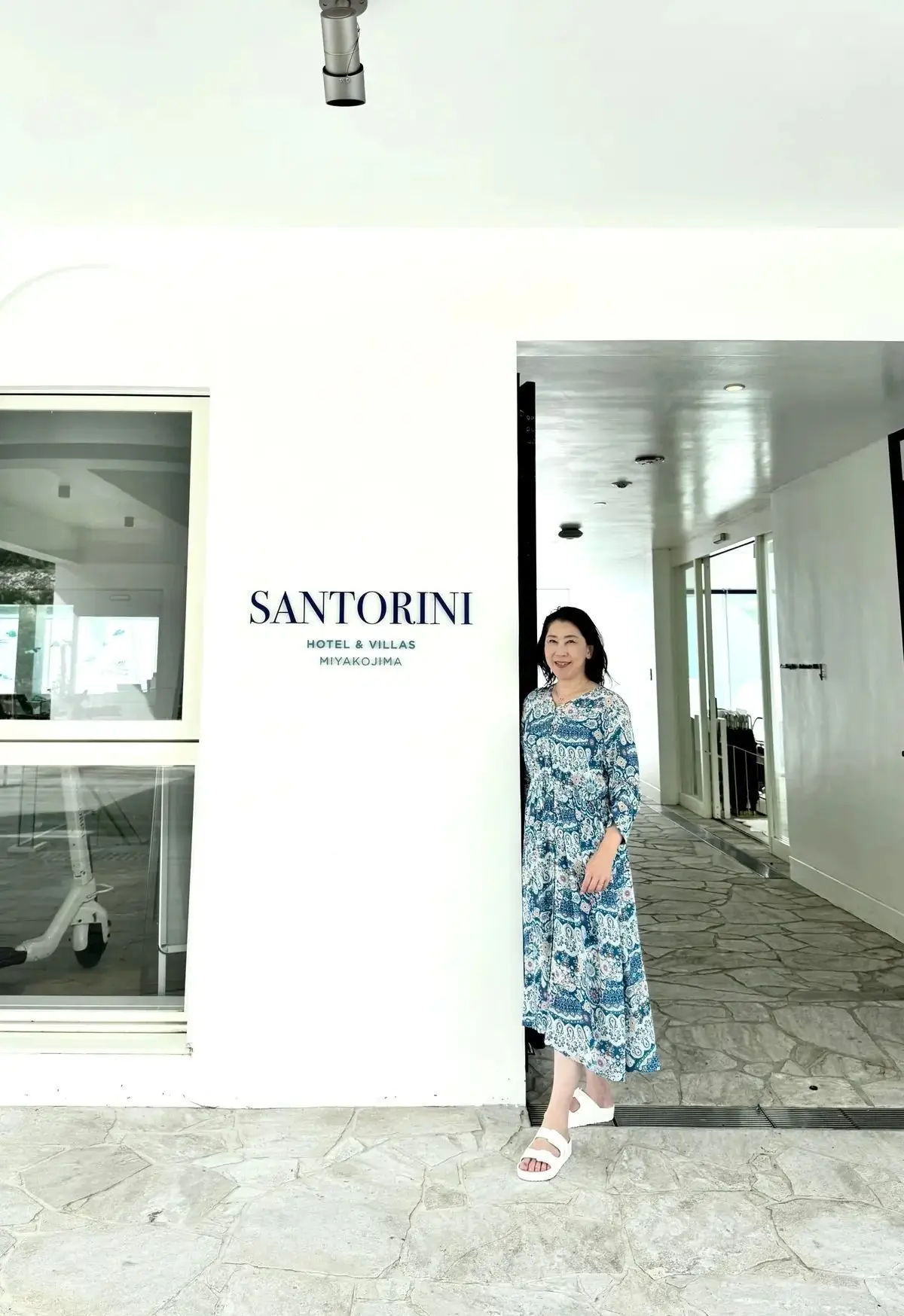 白い壁に「SANTORINI HOTEL & VILLAS MIYAKOJIMA」のロゴがある。女性が、青と白の柄のロングワンピースと白いサンダルを身につけ、笑顔で立っている。背景には、明るい廊下が映っている。