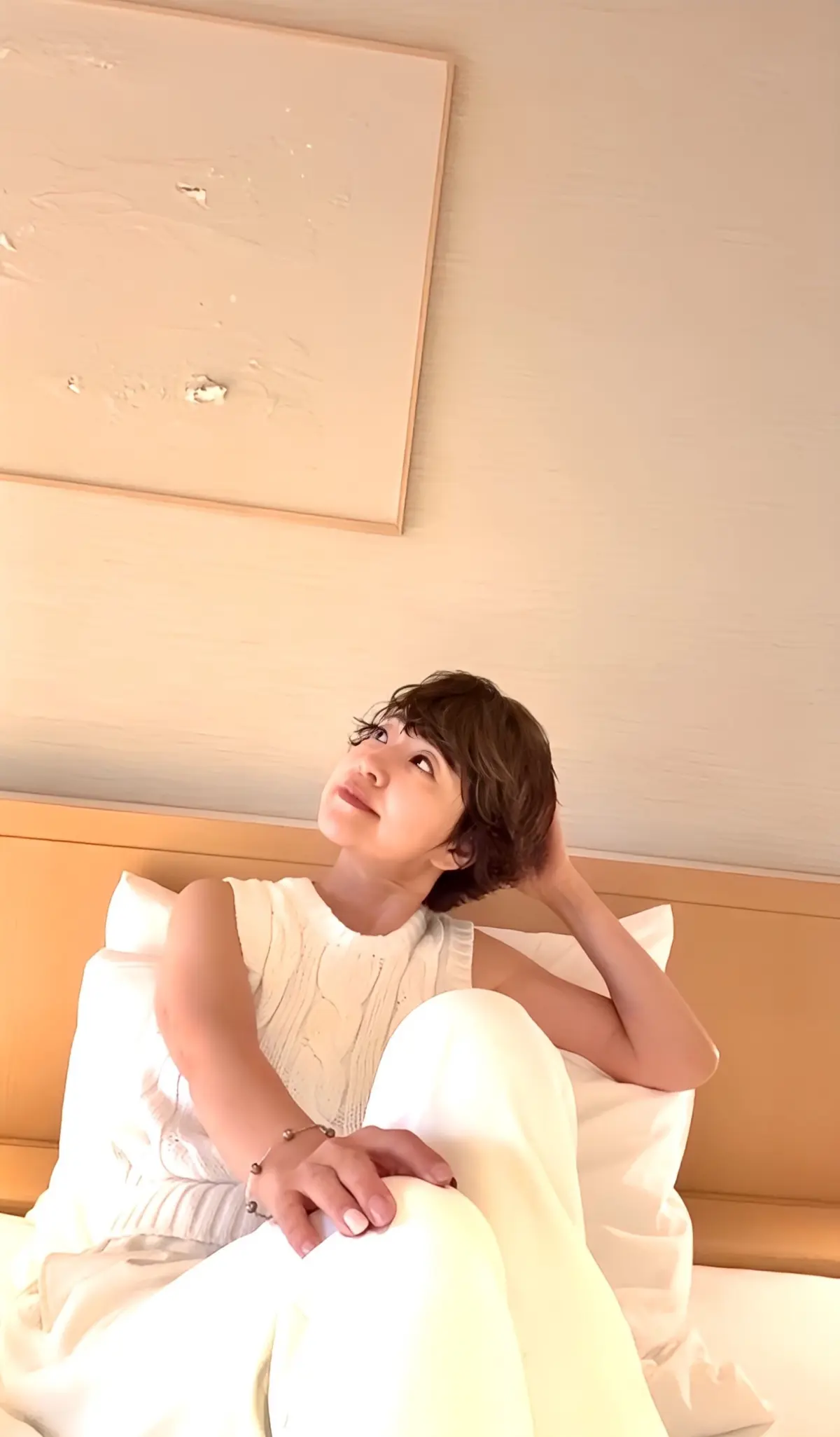 ベッドに座り、白い上下セットアップを着た女性。右手を頭に当て、微笑みながら上を見上げている。背景には淡いベージュの壁と、抽象画が飾られている。