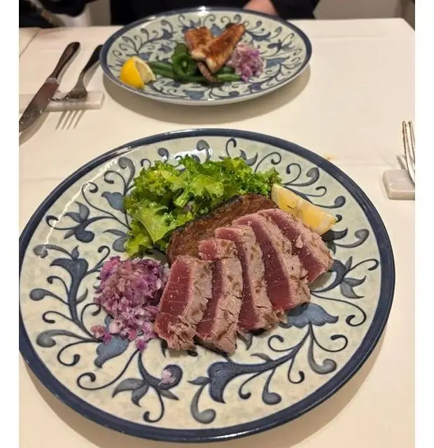 青い模様の皿に盛り付けられた、薄切りにした肉料理。レタス、紫色の玉ねぎ、レモンスライスが添えられている。背景には同じ皿に入った別の料理が見える。