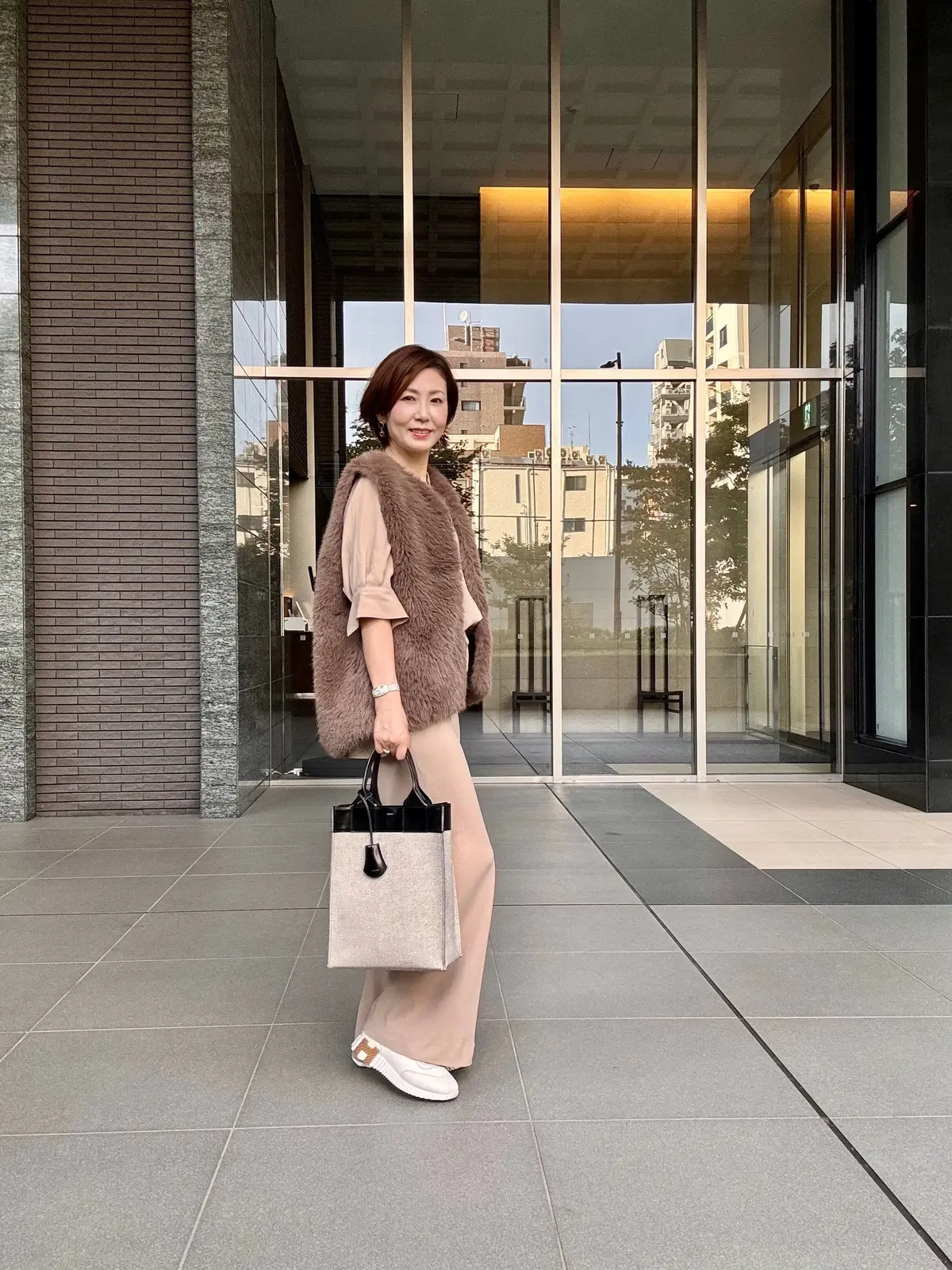 ベージュのパンツとタンクトップに茶色のファーベスト、白いスニーカーを着用した女性が立っている。背景はガラス張りの建物で、外の景色が映り込んでいる。右手に黒とベージュのトートバッグを持っている。